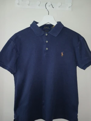 Mörkblå piké från Polo Ralph Lauren - Klassisk mörkblå pikétröja från Polo Ralph Lauren med korta ärmar och tre knappar framtill. Tröjan har den ikoniska broderade loggan på bröstet och är tillverkad i mjuk bomull. Perfekt till jeans eller shorts för en clean look.