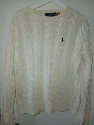 Vit kabelstickad tröja Polo Ralph Lauren - Kabelstickad vit tröja från Polo Ralph Lauren med rund halsringning och blå broderad logga på bröstet. Tröjan har långa ärmar och ribbade muddar vid ärmslut och nederkant. Klassisk och tidlös design i mjukt material.