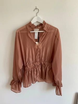 Rosa volangblus - Söt rosa blus från Vicolo med volangdetaljer vid krage och midja. Blusen har knappar framtill, transparent tyg och långa ärmar med volang vid ärmslut. Perfekt för en trendig och feminin look. Helt oanvänd.