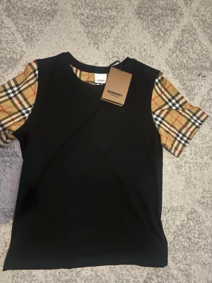 Svart t-shirt med Burberry-rutor - Snygg svart t-shirt från Burberry med klassiska beige, röda och svarta rutiga ärmar. T-shirten har rund halsringning och är tillverkad i mjukt material. Perfekt för dig som vill ha en stilren look med en twist. Storleken passar för M/L. Priset är ej hugget i sten!