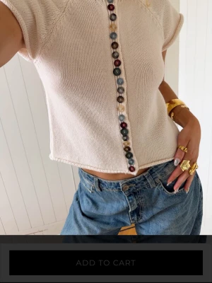 Ljusbeige stickad topp med knappar - Trendig ljusbeige stickad topp med korta ärmar och färgglada knappar framtill. Toppen är croppad och har en avslappnad passform som passar perfekt till jeans eller kjol. Snygg detalj med olika färger på knapparna som ger en unik look. Ifrån design by si o storlek M/L, nypris 590kr