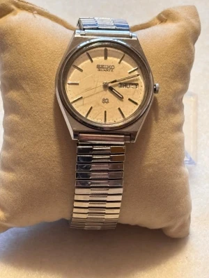 Seiko Quartz silverfärgad klocka - Klassisk Seiko Quartz klocka med rund, ljus urtavla och dag- och datumvisning. Boetten och armbandet är i silverfärgad metall med stilren, tidlös design. Länkat armband som ger en clean look. Perfekt för dig som gillar retrovibbar och enkelhet.