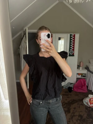 Svart volangblus - Snygg svart blus med volangprydda axlar och ryschdetaljer längs ärmöppningarna. Blusen har en rak passform och en liten v-ringning i nacken. Perfekt att styla med jeans eller kjol för en trendig look. Materialet känns lätt och luftigt.