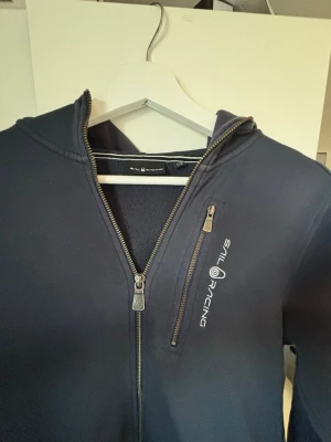 Mörkblå hoodie från Sail Racing - Snygg mörkblå hoodie från Sail Racing med dragkedja framtill och en extra bröstficka med zip. Loggan är tryckt i vitt på bröstet. Hoodien har långa ärmar och en klassisk huva. Perfekt för en avslappnad och sportig stil.