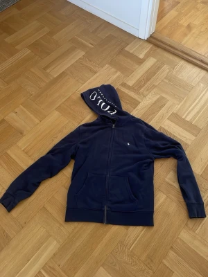 Marinblå hoodie från Polo Ralph Lauren -  marinblå hoodie från Polo Ralph Lauren med dragkedja framtill och stor huva med tydligt tryck. Klassisk logga på bröstet och två rymliga fickor. Tillverkad i mjukt material som känns skönt mot huden. Perfekt för en avslappnad och stilren look.
