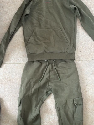 Olivgrön Tracksuit från McKenzie - Säljer en olivgrön hoodie från McKenzie med klassisk känguruficka framtill och ribbade muddar. Hoodien har en enkel och stilren design med diskret logga på bröstet. Tillverkad i mjukt bomullsmaterial som känns skönt mot huden.