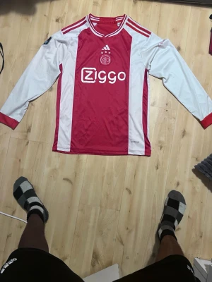 Ajax långärmad matchtröja Adidas - Säljer en långärmad Ajax Amsterdam matchtröja från Adidas i rött och vitt med Ziggo sponsortryck på bröstet. Klassisk design med klubbmärke och tre stjärnor ovanför loggan. Perfekt för dig som älskar fotboll och vill representera Ajax.