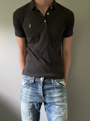 Svart pikétröja från Ralph Lauren - Klassisk svart pikétröja från Ralph Lauren med korta ärmar och liten broderad logga på bröstet. Tröjan har krage och knappar vid halsen. Perfekt till jeans eller shorts för en avslappnad men snygg look.