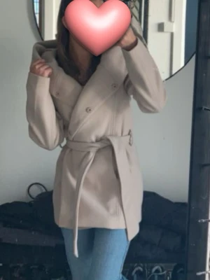 Beige kappa med knytskärp - Stilren beige kappa med dubbelknäppning och brett slag. Kappan har ett matchande knytskärp i midjan och är tillverkad i ett mjukt tyg som ger en snygg siluett. Perfekt för dig som gillar klassisk och enkel stil.