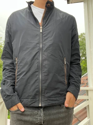 Massimo Dutti jacka - Snygg mörkblå vindjacka med ståkrage och dragkedja framtill. Jackan har två framfickor med kontrasterande dragkedjor och en extra ficka på ärmen. Tillverkad i lätt och vindtätt material, perfekt för blåsiga dagar. Riktigt snygga detaljer på jackan som ger den ett rejält lyft.