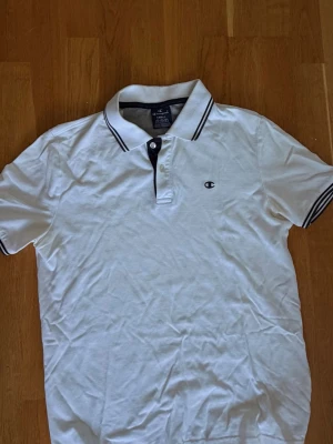 Vit pikétröja från Champion - Vit pikétröja från Champion med svart broderad logga på bröstet. Tröjan har klassisk krage med svarta ränder och knappar framtill. Storlek S