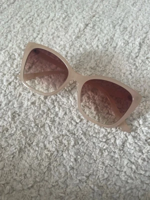 Beige cat eye solglasögon - Snygga solglasögon i cat eye-modell med beige bågar och bruna tonade glas. Ramen är i plast och ger en trendig och modern look. Perfekta för soliga dagar.
