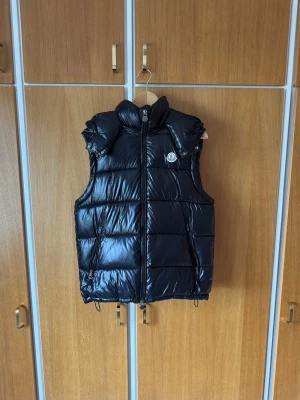 Svart dunväst från Moncler - Snygg svart dunväst från Moncler med glansig finish och luva som är avtagbar. Västen har dragkedja framtill, två sidofickor och Moncler-logga på bröstet. Perfekt för lager-på-lager och ger en sportig vibe till din outfit.