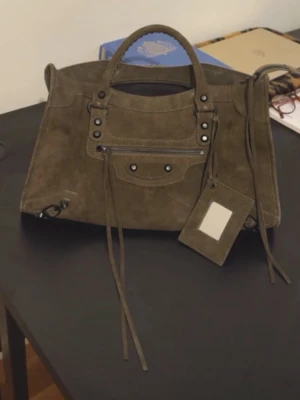 Olivgrön mocka handväska med detaljer - (KOMMER MED SIN STRAP OCH SPEGEL!) Snygg olivgrön handväska i mocka med dekorativa nitar och dragkedja framtill. Väskan har dubbla handtag, långa mockaband och en matchande tag. Perfekt för dig som gillar en cool och avslappnad stil.