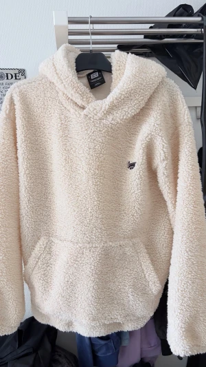 Beige teddy hoodie från Urban Classics - Mysig beige teddy hoodie från Urban Classics med stor huva och magficka. Tillverkad i fluffigt, mjukt material som ger en riktigt chill vibe. Liten logga broderad på bröstet. Perfekt för dig som vill ha något både bekvämt och snyggt.