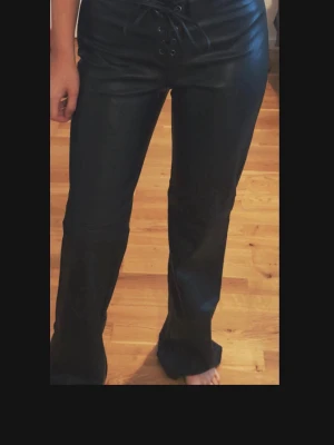 Svarta low waist byxor - Snygga svarta byxor i skinnimitation med utsvängda ben och snörning framtill i midjan. Byxorna har en tight passform upptill och sitter snyggt över höften. Perfekta för dig som vill sticka ut med en trendig look.