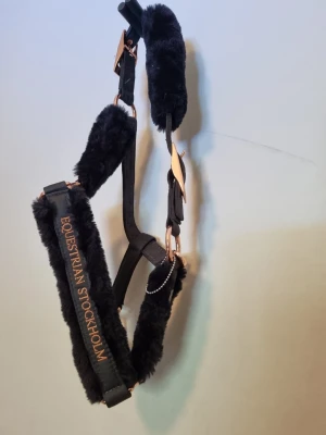 Svart grimma från Equestrian Stockholm - Säljer en svart hästgrimma från Equestrian Stockholm med mjuk fuskpäls och detaljer i roséguld. Grimman har broderad logga och justerbara spännen. Perfekt för dig som vill ge din häst extra komfort och stil. Dark sky heter färgen. Den är aldrig använd. Testade att ta på den på min häst men storleken var för liten. 