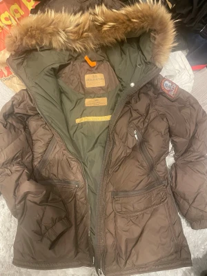 Brun dunjacka från Parajumpers - Snygg brun dunjacka från Parajumpers med stor huva och avtagbar fuskpäls. Jackan har flera fickor med dragkedja och patch på ärmen. Insidan är olivgrön och jackan har en klassisk, puffig passform. Perfekt för kalla vinterdagar.