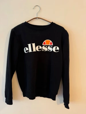 Svart sweatshirt från Ellesse - Säljer en svart sweatshirt från Ellesse med stor vit logga och orange detalj på bröstet. Tröjan har rund halsringning, långa ärmar och ribbade muddar vid ärmslut och nederkant. Perfekt för en avslappnad och sportig stil.