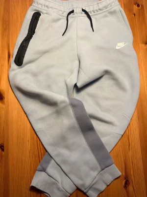Ljusblå mjukisbyxor från Nike - Snygga ljusblå mjukisbyxor från Nike med svart dragsko i midjan och vit logga på vänster sida. Byxorna har en svart dragkedjeficka på höger sida och ribbade muddar vid bensluten. Perfekta för chill dagar och streetwear.