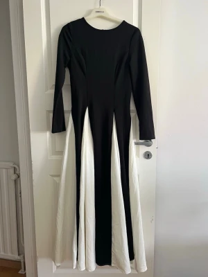 Svartvit långklänning med långa ärmar - Snygg långklänning i svart och vitt med långa ärmar och rund halsringning. Klänningen har en elegant, figurnära passform upptill och vida paneler i kjolen som ger en cool kontrast mellan svart och vitt. Perfekt för dig som vill sticka ut.