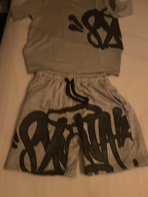Grått set från Syna world  med graffiti-tryck - Säljer ett grått set från Stay med matchande t-shirt och shorts. Båda plaggen har stora svarta graffiti-inspirerade tryck framtill. Materialet är mjukt och bekvämt, perfekt för en avslappnad stil. Elastisk midja på shortsen för extra komfort.