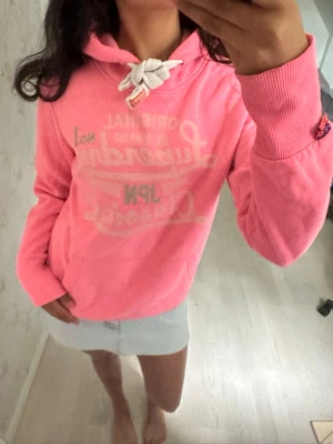 Rosa hoodie med tryck och huva - Säljer en rosa hoodie med vit text på bröstet och stor huva med vita snören. Hoodien har ribbade muddar vid ärmarna och en klassisk känguruficka framtill. Perfekt för en avslappnad och trendig look.