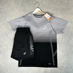 Grå och svart träningsset från Asics - Säljer ett snyggt träningsset från Asics med en grå och svart t-shirt och matchande svarta shorts. T-shirten har ett diskret mönster och Asics-logga på bröstet, medan shortsen har logga och grafiskt tryck på sidan. Perfekt för löpning eller gym.