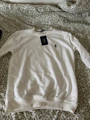 Vit sweatshirt från Polo Ralph Lauren - Klassisk vit sweatshirt från Polo Ralph Lauren med rund halsringning och broderad logga på bröstet. Tröjan har ribbade muddar vid ärmslut och nederkant. Perfekt för en clean och stilren look.