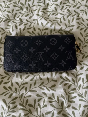Svart plånbok med monogram - Snygg svart plånbok med klassiskt monogrammönster i grått. Plånboken har dragkedja i guldton och flera fack för kort, sedlar och mynt. Perfekt för dig som vill ha både stil och funktion i vardagen.