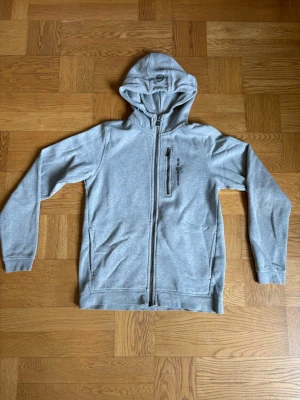 Sail racing hoddie - Snygg grå hoodie från Sail Racing med dragkedja framtill och en praktisk bröstficka med dragkedja. Tillverkad i mjukt bomullsmaterial och har ribbade muddar vid ärmslut och nederkant. Perfekt för en avslappnad stil och enkel att matcha.Finss några små fläckar på den