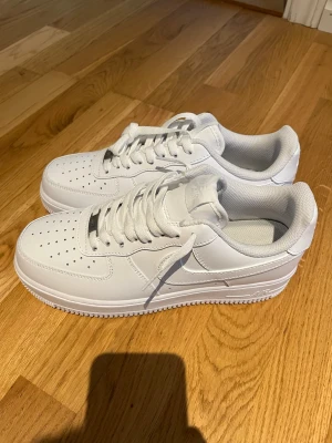 Nike Air Force 1 vita sneakers - Klassiska helvita Nike Air Force 1 sneakers i lågt utförande. Skorna har ovandel i slätt läder, perforeringar på tån för ventilation och den ikoniska swoosh-loggan på sidan. Tjock vit sula och vita skosnören ger en clean och tidlös look.