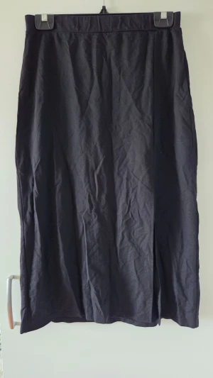 Svart långkjol med slits - En enkel och stilren svart långkjol. Kjolen har rak passform, är knälång, och har en diskret slits framtill för extra rörelsefrihet. Perfekt att matcha med en crop top eller stickad tröja för en clean look.