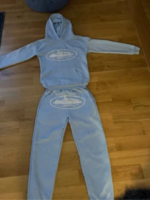 Ljusblått corteiz med tryck - Säljer ett ljusblått corteiz hoodie och matchande byxor. Setet har ett vitt tryck med motiv på både tröjan och byxorna. Hoodien har magficka och byxorna har resår i midjan. Perfekt för chill dagar hemma.
