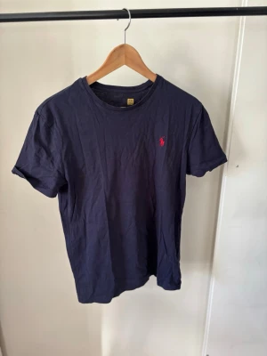 Mörkblå t-shirt från Polo Raulph Lauren - Snygg mörkblå t-shirt i bomull med klassisk rund hals och korta ärmar. På bröstet finns en liten röd broderad logga som ger en stilren touch. Perfekt till jeans eller shorts för en enkel och clean look.