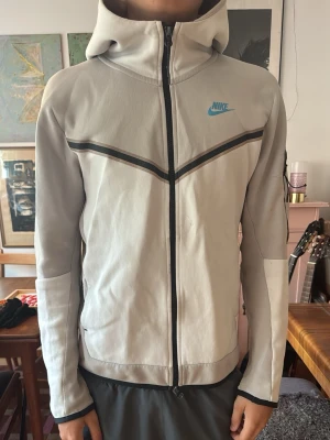 Nike Tech Fleece hoodie ljusgrå - Nike Tech Fleece hoodie i ljusgrått med svart dragkedja och svarta detaljer över bröstet. Har huva och Nike-logga i blått på vänster sida. Tillverkad i mjukt och lätt material som passar perfekt för en sportig och avslappnad stil. Köpt på loppas så ingen äkthet garanteras 