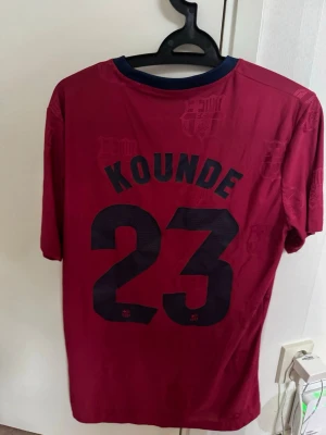 Vinröd fotbollströja Kounde 23 - Snygg vinröd fotbollströja med mörkblå detaljer och trycket 'KOUNDE 23' på ryggen. Tröjan har korta ärmar och rund halsringning med kontrastfärg. Perfekt för dig som vill stötta ditt favoritlag på match eller träning.