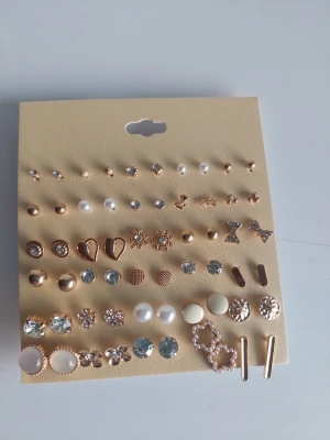 Set med 30 par örhängen i guld och pärlor - Säljer ett set med 30 par örhängen i olika former och stilar. Färgerna är främst guld, vitt och transparent. Innehåller bland annat pärlor, strass, hjärtan, stjärnor, rosetter och enkla studs. Materialet är metall, plast och akryl. Perfekt för att mixa och matcha till olika looks.