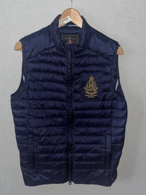 Morris väst  - Snygg marinblå dunväst från Morris med guldfärgat broderat emblem på bröstet. Västen har horisontella quiltade ränder, ståkrage och dragkedja framtill. Perfekt för lager-på-lager och har en sportig, klassisk look. 15. (Västen är ej så glansig som på bild utan det är pga att det är fotat med blixt)