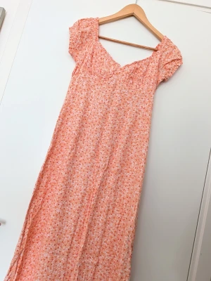 H&M lång blommig sommarklänning i orange - Söt långklänning i ljus orange med små vita blommor och korta puffärmar. Klänningen har smock upptill och en lätt v-ringad halsringning. Perfekt för varma dagar och har en luftig, flowy passform. Storlek 40. Från viskos. Som ny. 