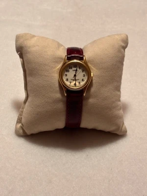 Lorus Klassisk guldklocka med vinrött läderband - Stilren klocka med rund vit urtavla, svarta siffror och guldfärgat boett. Armbandet är i vinrött läder med klassiskt spänne. Perfekt accessoar för dig som gillar tidlös design och eleganta detaljer.