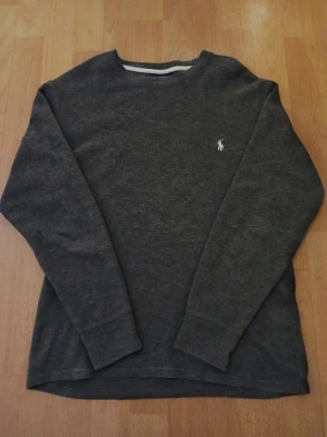 Mörkgrå stickad tröja från Polo Ralph Lauren - Säljer en mörkgrå stickad tröja från Polo Ralph Lauren med klassisk rund hals och långärmad passform. Tröjan har en vit broderad logga på bröstet och är tillverkad i mjukt stickat material som är perfekt för kyliga dagar. Axelbredd: 48cm / Längd: 62cm. Nypris: 995kr / Mitt pris: 449kr