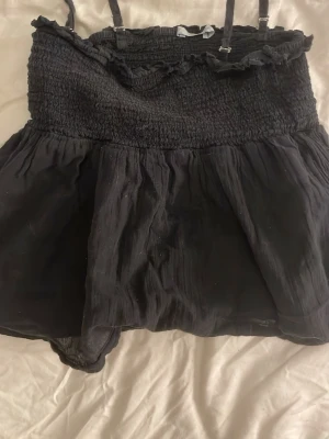 Svart ärmlös linnetopp med volang - Snygg svart linnetopp med smockad överdel och tunna axelband. Toppen har volangdetalj upptill och ett flowy, lätt tyg nedtill som ger en luftig känsla. Perfekt för varma dagar och enkel att styla med jeans eller kjol.