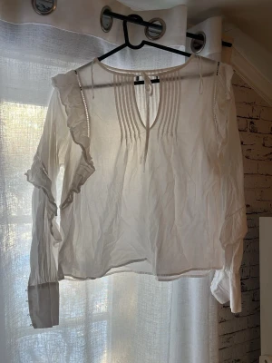 Vit blus - Söt vit blus med långa ärmar, volangdetaljer längs axlarna och knytband vid halsen. Blusen har dekorativa veck framtill och breda manschetter med knappar. Perfekt för en trendig och romantisk look.