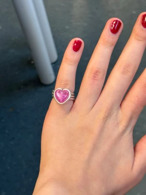 Silverfärgad ring med rosa hjärta - Unik ring i silverfärgad metall med ett stort rosa hjärta i mitten. Ringen har en cool, spiralformad design och hjärtat har en glansig yta som verkligen poppar. Perfekt för dig som vill ha något som sticker ut och ger en söt touch till din stil.
