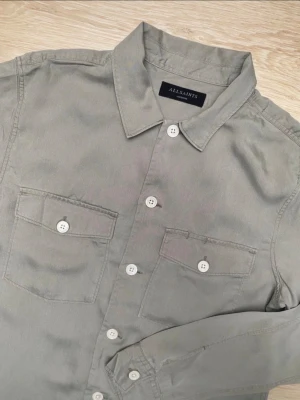 Grå overshirt från AllSaints - Stilren grå overshirt från AllSaints med klassisk krage, vita knappar och två bröstfickor med lock. Tillverkad i ett mjukt bomullstyg och har långa ärmar med knapp vid ärmslut. Perfekt lager-på-lager-plagg för en avslappnad look.