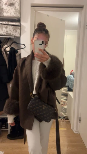 Brun kappa med pälskant och skärp - Snygg brun kappa med bred krage, pälskantade ärmslut och tillhörande skärp i midjan. Modellen är rak och oversize, vilket ger en trendig och avslappnad look.