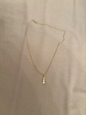 Guldhalsband med tre små pärlor - Snyggt och stilrent halsband i guldton med en hängande rad av tre vita pärlor. Kedjan är tunn och elegant, perfekt för att ge en subtil touch till din outfit. Passar dig som gillar minimalistiska smycken med en klassisk twist.