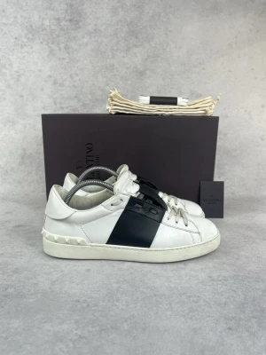 Valentino vita sneakers med svart detalj - Snygga vita sneakers från Valentino med bred svart läderdetalj över sidan. Skorna har klassisk låg profil, vita snören och nitar på hälen. Tillverkade i läder med en chunky sula som ger en modern vibe.