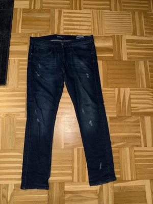 Mörkblå slitna slim-fit jeans från Blend - Säljer ett par mörkblå jeans från Blend med snygga slitningar och tvättade detaljer. Klassisk femficksmodell, slim-fit och knappgylf. Jeansen är gjorda i bomull med lite stretch för extra komfort.